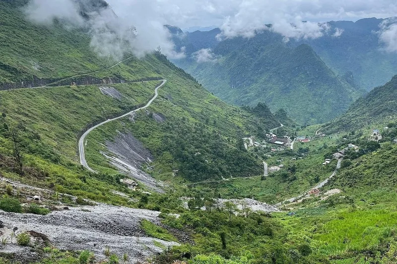 A winding mountain road descends through misty green peaks into a small village in Ha Giang.