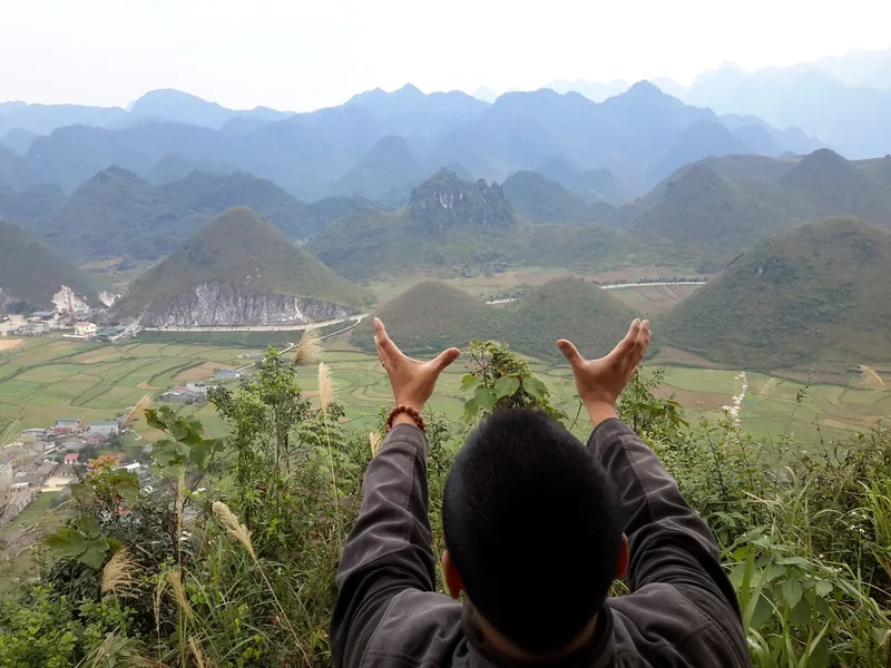 A traveler raises his hands to appear to hold twin peaks above the valley of Ha Giang's karst mountains.