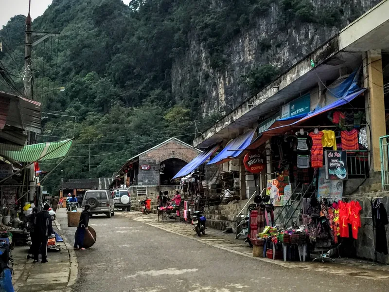 A quiet street in Dong Van, Ha Giang lined with shops selling clothing beneath a towering limestone cliff.