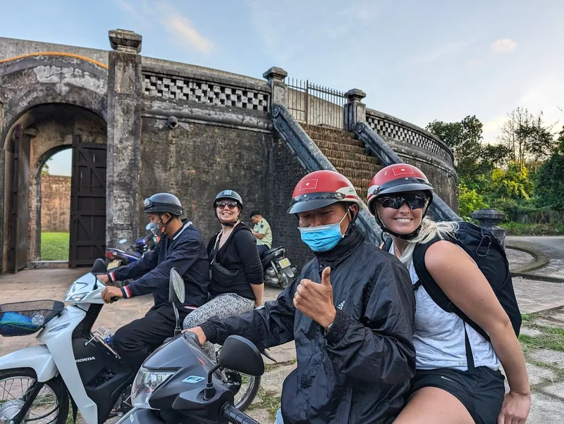 Tourists on motorbikes with local guides pause beside an old stone gate at the Hue citadel walls.