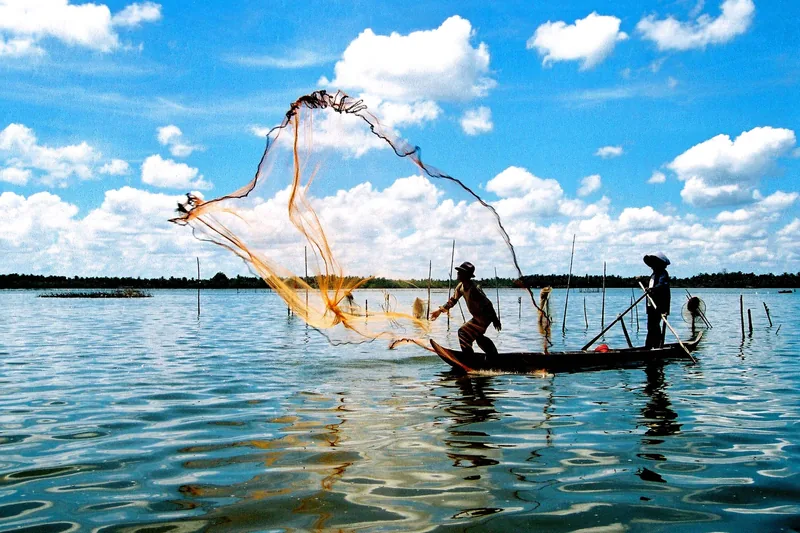 A fisherman casts a wide net from a wooden boat on Tam Giang Lagoon under a bright blue sky.