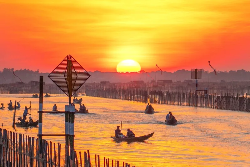 Fishing boats glide across Tam Giang Lagoon under a vivid orange sunset, framed by bamboo fences.