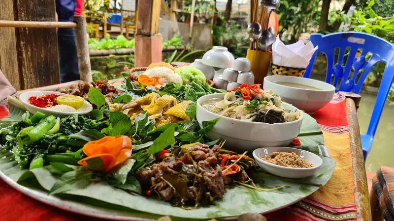 A large tray of traditional Mai Chau dishes with grilled meats, herbs, vegetables, and soup served outdoors. A large tray of traditional Mai Chau dishes with grilled meats, herbs, vegetables, and soup served outdoors.