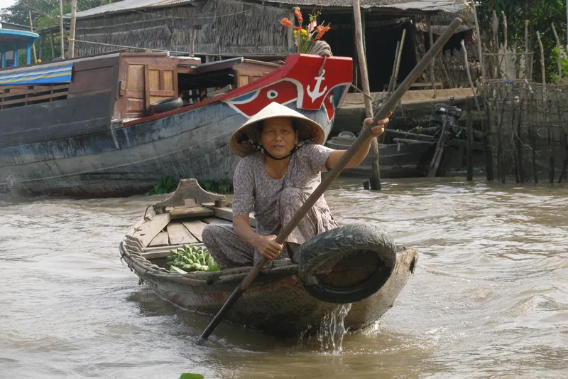 A woman in a conical hat rows a small wooden boat carrying produce at a floating market in the Mekong Delta.