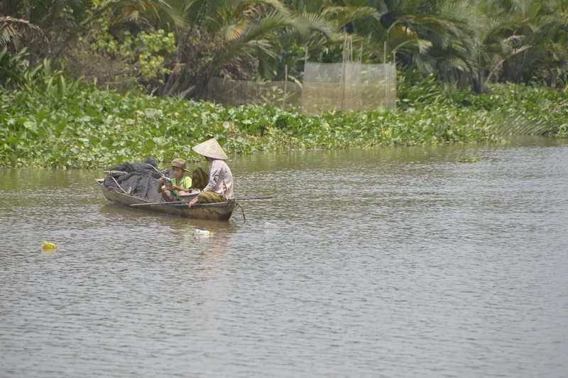 A wooden boat carrying two people in conical hats drifts along a calm river in the Mekong Delta.