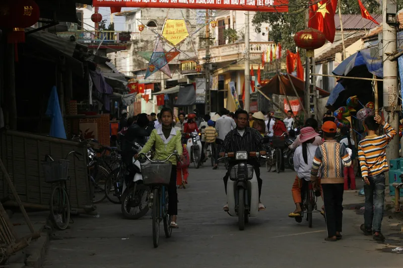 A busy Vietnamese village street decorated with red flags and lanterns during Tet celebrations, filled with cyclists and.
